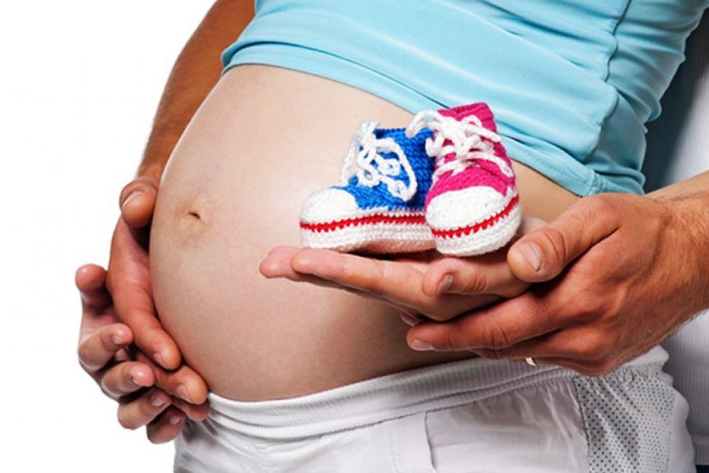 Беременная женщина держит на ладони обувь голубого и розового цвета